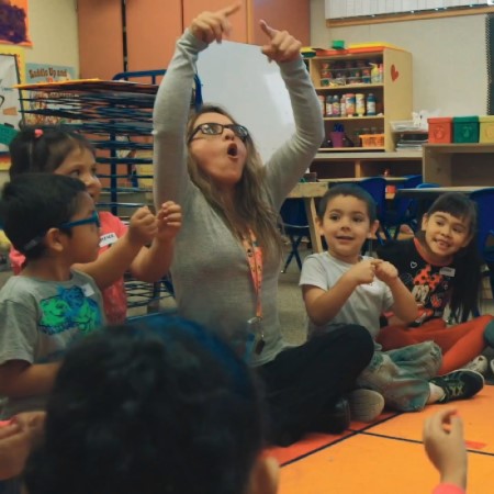 teacher gesturing above head, students look on and smile many copying the gesture